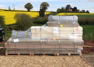 Pallets of wrapped lumber stacked on a pallet beside a rural road, with yellow rapeseed fields in the background.