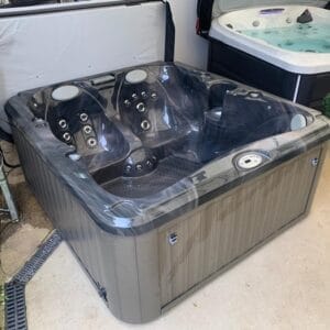 Gray outdoor hot tub with multiple seats and built-in jets on a concrete floor, in a showroom setting.
